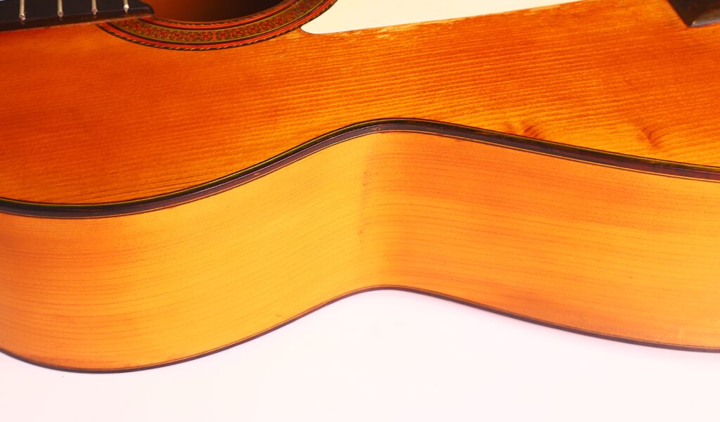 Nahaufnahme der Seite und der Oberseite einer Jose Ramirez 1955 Flamenco-Gitarre, die die Holzmaserung, den gewölbten Korpus und einen Teil des Schalllochs vor einem weißen Hintergrund zeigt. Nahaufnahme der Seite und der Oberseite einer Jose Ramirez 1955 Flamenco-Gitarre, die die Holzmaserung, den gewölbten Korpus und einen Teil des Schalllochs vor einem weißen Hintergrund zeigt.