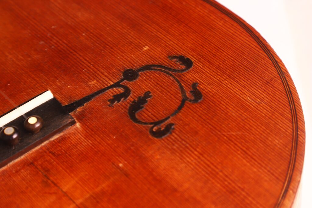 Nahaufnahme des Holzkörpers der „Italienischen Romantikgitarre 1860“ mit einer detaillierten schwarzen Verzierung im Bereich des Stegs. Nahaufnahme des Holzkörpers der „Italienischen Romantikgitarre 1860“ mit einer detaillierten schwarzen Verzierung im Bereich des Stegs.