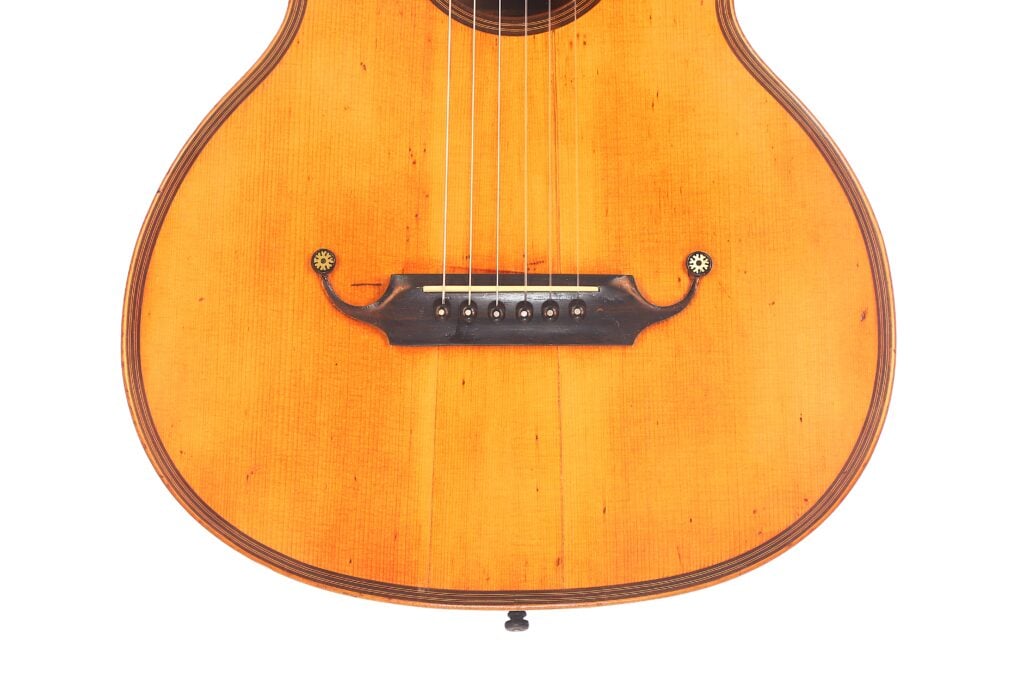 Nahaufnahme des Korpus der Markneukirchener Romantikgitarre von 1880, die den Steg, die Saiten und den unteren Teil der Decke vor weißem Hintergrund zeigt. Nahaufnahme des Korpus der Markneukirchener Romantikgitarre von 1880, die den Steg, die Saiten und den unteren Teil der Decke vor weißem Hintergrund zeigt.