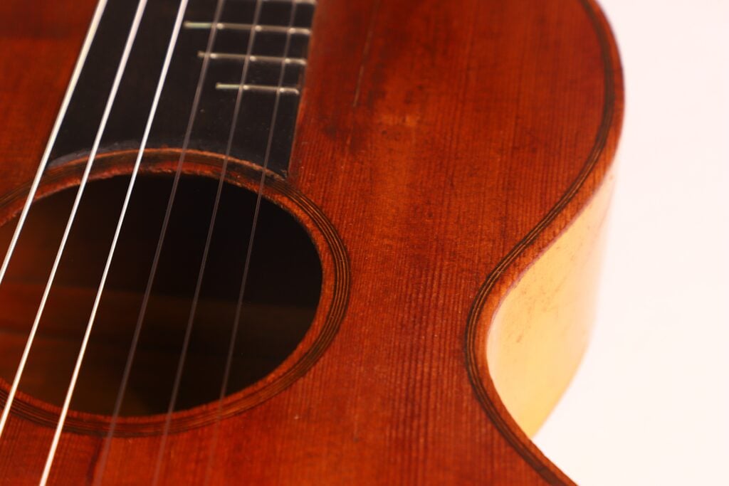 Nahaufnahme der „Italienischen Romantikgitarre 1860“ mit Fokus auf das Schallloch, die Saiten und den warmbraunen Holzkorpus vor einem hellen Hintergrund. Nahaufnahme der „Italienischen Romantikgitarre 1860“ mit Fokus auf das Schallloch, die Saiten und den warmbraunen Holzkorpus vor einem hellen Hintergrund.