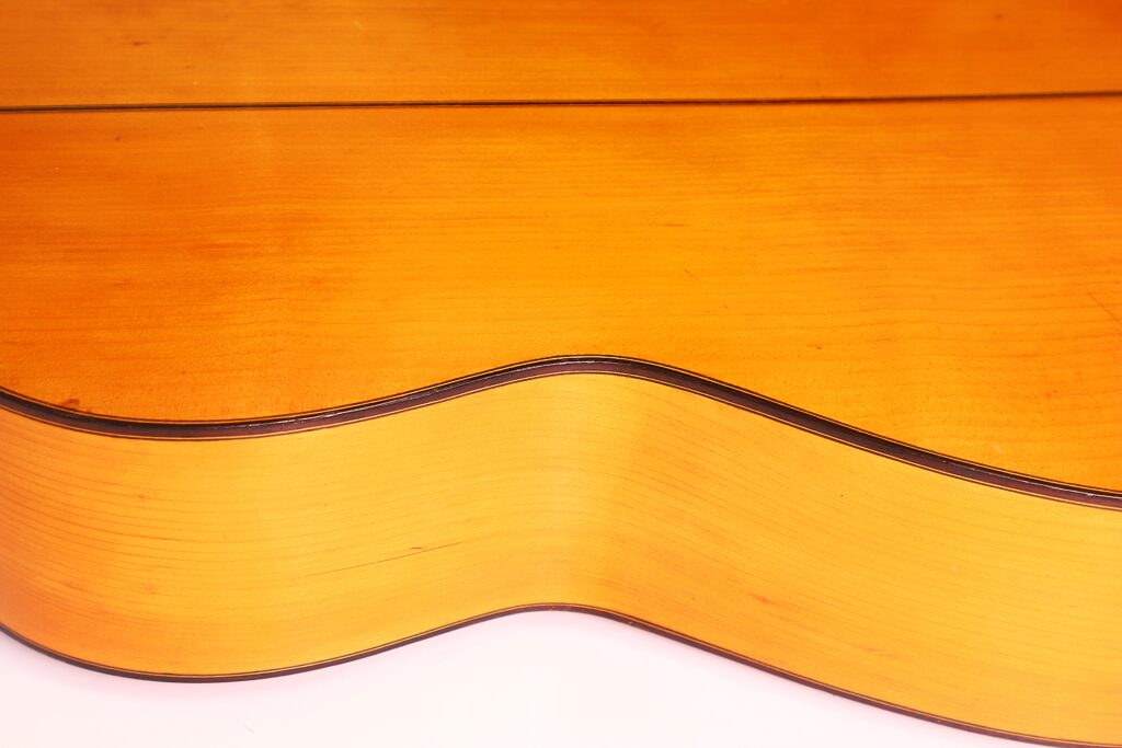 Nahaufnahme der Seite und des Korpus der Jose Ramirez 1955 Flamenco Gitarre. Hervorgehoben werden die Holzmaserung, die geschwungene Form und die dunklen Zierleisten mit Fokus auf die Kante der Gitarre vor einem weißen Hintergrund. Nahaufnahme der Seite und des Korpus der Jose Ramirez 1955 Flamenco Gitarre. Hervorgehoben werden die Holzmaserung, die geschwungene Form und die dunklen Zierleisten mit Fokus auf die Kante der Gitarre vor einem weißen Hintergrund.