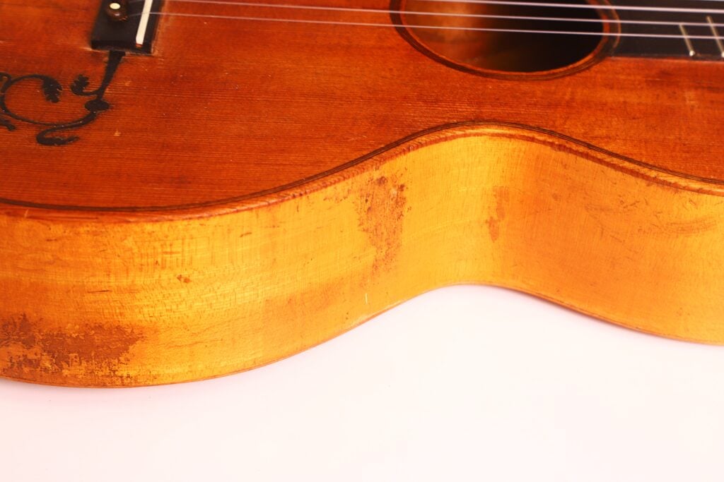 Nahaufnahme der Seite und des Korpus einer italienischen Romantikgitarre von 1860, die die Maserung des Holzes, Gebrauchsspuren und einen Teil des Schalllochs vor weißem Hintergrund hervorhebt. Nahaufnahme der Seite und des Korpus einer italienischen Romantikgitarre von 1860, die die Maserung des Holzes, Gebrauchsspuren und einen Teil des Schalllochs vor weißem Hintergrund hervorhebt.