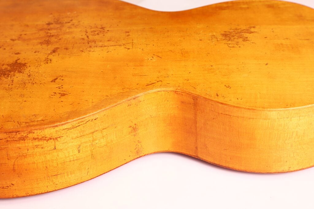 Nahaufnahme der Rückseite und der Seite der „Italienischen Romantikgitarre 1860“, die die geschwungene Holzform sowie sichtbare Kratzer und Abnutzungsspuren auf der Oberfläche hervorhebt. Nahaufnahme der Rückseite und der Seite der „Italienischen Romantikgitarre 1860“, die die geschwungene Holzform sowie sichtbare Kratzer und Abnutzungsspuren auf der Oberfläche hervorhebt.