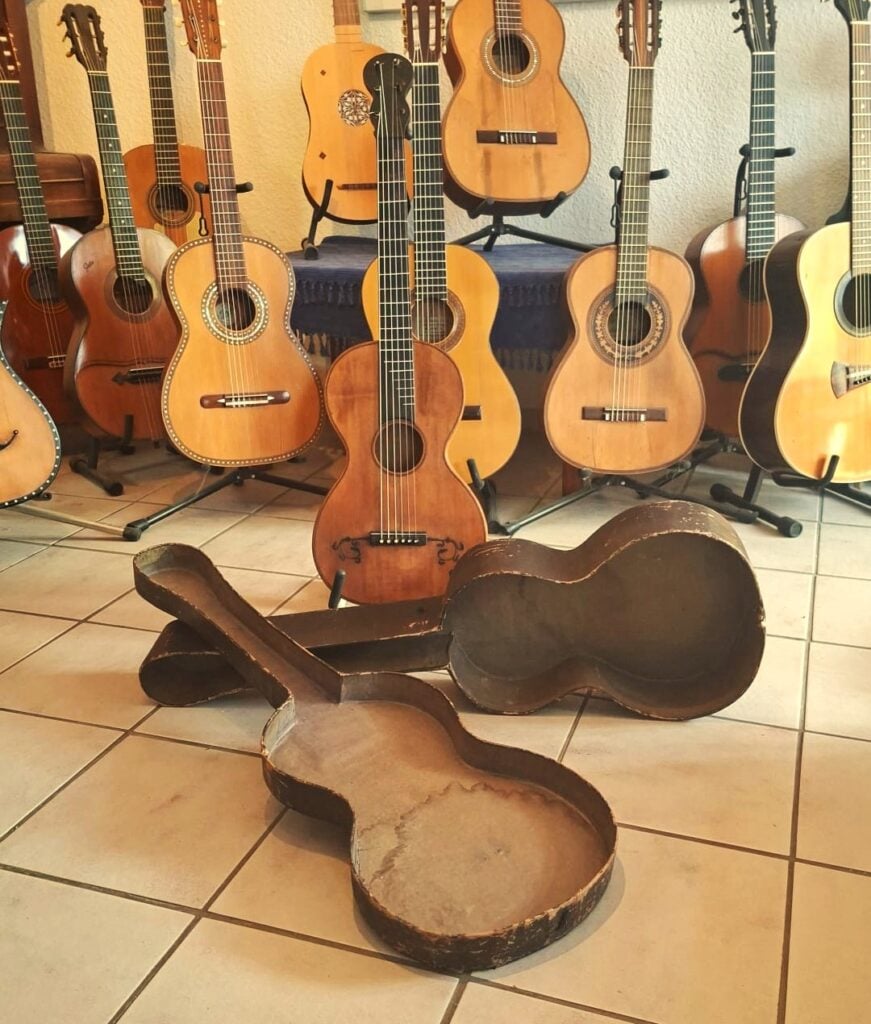 Eine italienische Romantikgitarre von 1860 steht aufrecht neben anderen Akustikgitarren an einer Wand. Im Vordergrund liegt ein offener, alter Gitarrenkoffer leer auf dem Fliesenboden. Eine italienische Romantikgitarre von 1860 steht aufrecht neben anderen Akustikgitarren an einer Wand. Im Vordergrund liegt ein offener, alter Gitarrenkoffer leer auf dem Fliesenboden.