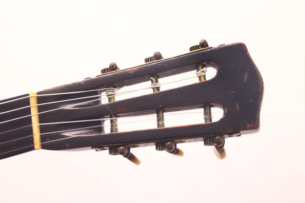 Nahaufnahme der Kopfplatte der Romantikgitarre Markneukirchen 1880 mit Nylonsaiten, Stimmwirbeln und einer leicht abgenutzten dunklen Holzoberfläche vor einem schlichten hellen Hintergrund. Nahaufnahme der Kopfplatte der Romantikgitarre Markneukirchen 1880 mit Nylonsaiten, Stimmwirbeln und einer leicht abgenutzten dunklen Holzoberfläche vor einem schlichten hellen Hintergrund.