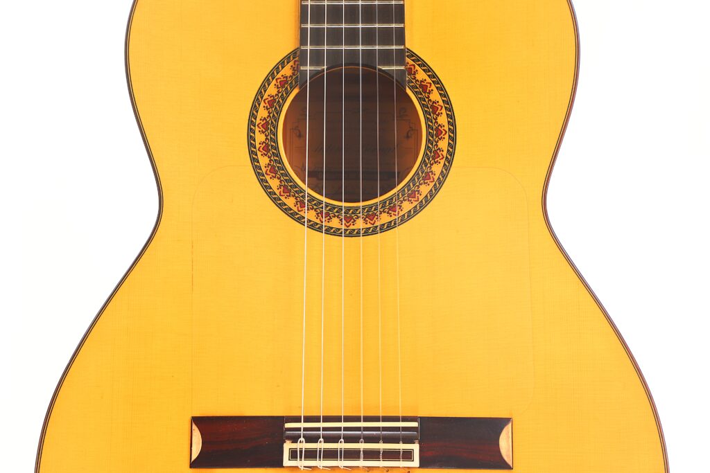 Eine Nahaufnahme des Korpus einer Flamenco-Gitarre von Antonio Bernal aus dem Jahr 2009 mit sechs Saiten, einem verzierten Schallloch und einem Teil des Griffbretts vor weißem Hintergrund. Eine Nahaufnahme des Korpus einer Flamenco-Gitarre von Antonio Bernal aus dem Jahr 2009 mit sechs Saiten, einem verzierten Schallloch und einem Teil des Griffbretts vor weißem Hintergrund.