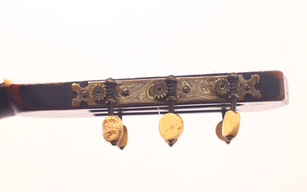 Nahaufnahme der Stimmwirbel und der dekorativen Metallplatte an der Kopfplatte der Romantikgitarre Markneukirchen 1880 vor einem sanft verschwommenen Hintergrund. Nahaufnahme der Stimmwirbel und der dekorativen Metallplatte an der Kopfplatte der Romantikgitarre Markneukirchen 1880 vor einem sanft verschwommenen Hintergrund.