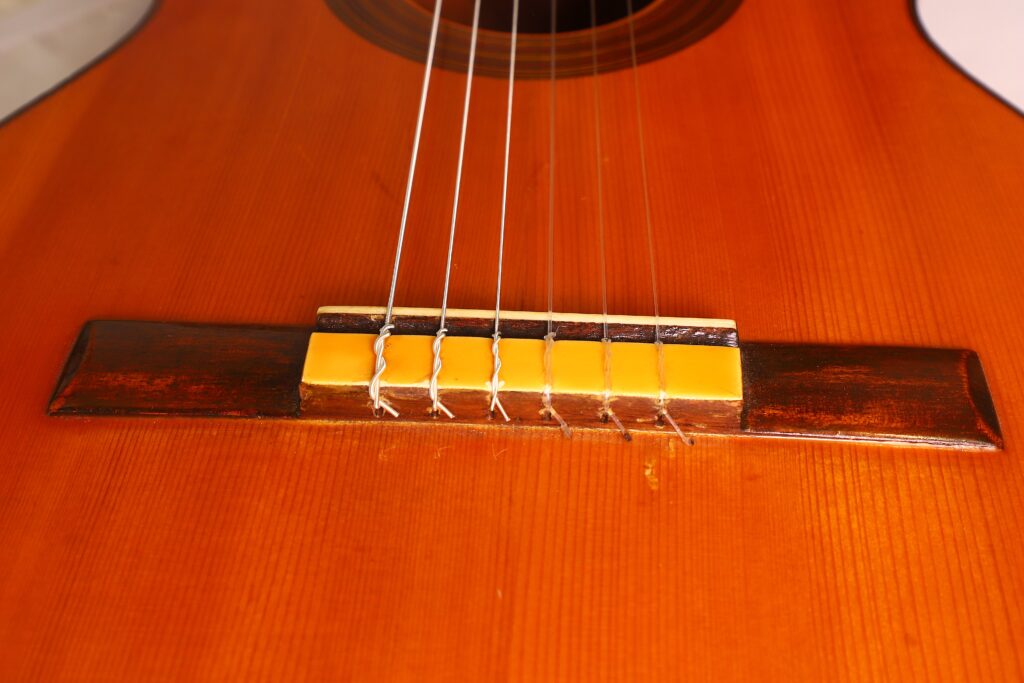 Nahaufnahme des Stegs der Ricardo Sanchis Nacher 1940 Flamenco Gitarre. Man sieht sechs Saiten, die am hölzernen Steg des Gitarrenkorpus befestigt sind, mit einer warmen braunen und gelben Lackierung. Nahaufnahme des Stegs der Ricardo Sanchis Nacher 1940 Flamenco Gitarre. Man sieht sechs Saiten, die am hölzernen Steg des Gitarrenkorpus befestigt sind, mit einer warmen braunen und gelben Lackierung.
