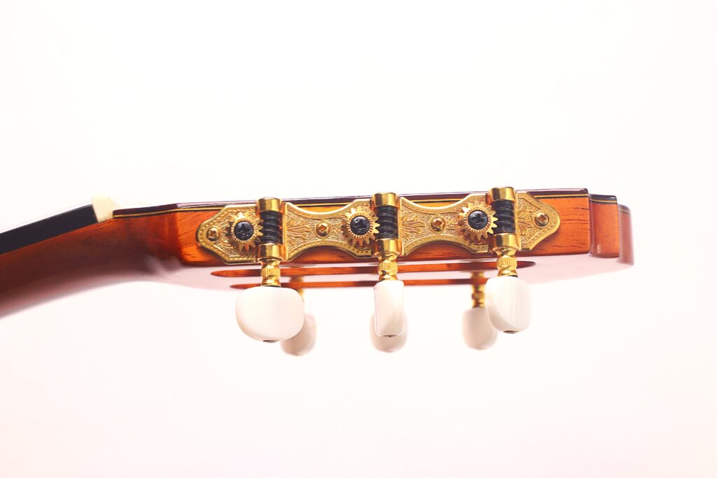 Nahaufnahme der Stimmwirbel und des Kopfes einer Antonio Bernal Flamenco-Gitarre aus dem Jahr 2009, mit drei weißen Stimmknöpfen auf einem verzierten goldenen Mechanismus vor einem schlichten weißen Hintergrund. Nahaufnahme der Stimmwirbel und des Kopfes einer Antonio Bernal Flamenco-Gitarre aus dem Jahr 2009, mit drei weißen Stimmknöpfen auf einem verzierten goldenen Mechanismus vor einem schlichten weißen Hintergrund.