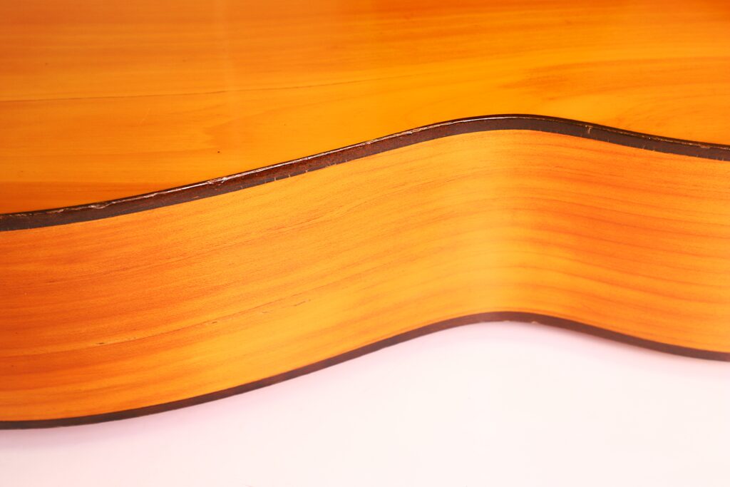 Nahaufnahme der Ricardo Sanchis Nacher 1940 Flamenco-Gitarre, die den glänzenden, geschwungenen Holzkorpus mit dunkler Verzierung entlang der Kante vor einem weißen Hintergrund zeigt. Nahaufnahme der Ricardo Sanchis Nacher 1940 Flamenco-Gitarre, die den glänzenden, geschwungenen Holzkorpus mit dunkler Verzierung entlang der Kante vor einem weißen Hintergrund zeigt.