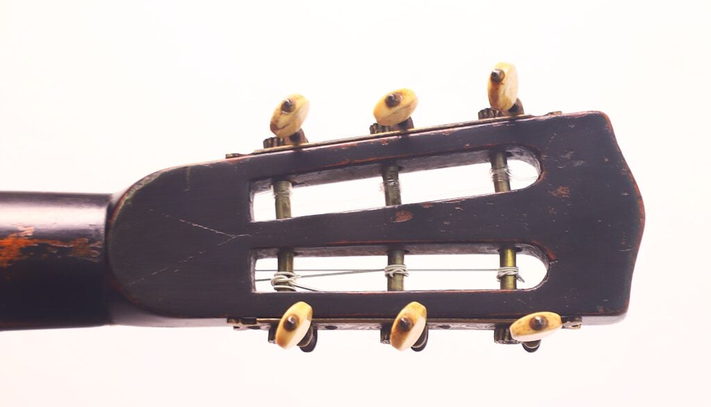 Nahaufnahme der Kopfplatte der Markneukirchener Romantikgitarre von 1880 mit sechs Stimmwirbeln, abgenutztem Holz und um die Wirbel gewickelten Metallsaiten vor weißem Hintergrund. Nahaufnahme der Kopfplatte der Markneukirchener Romantikgitarre von 1880 mit sechs Stimmwirbeln, abgenutztem Holz und um die Wirbel gewickelten Metallsaiten vor weißem Hintergrund.