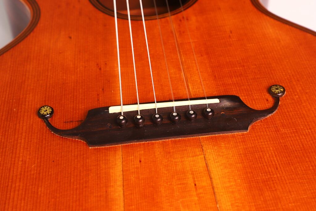 Nahaufnahme des Stegs und der Saiten der Romantikgitarre Markneukirchen 1880. Hervorzuheben sind die feine Holzmaserung und die verzierten Stimmknöpfe mit einer warmen, rotbraunen Lackierung. Nahaufnahme des Stegs und der Saiten der Romantikgitarre Markneukirchen 1880. Hervorzuheben sind die feine Holzmaserung und die verzierten Stimmknöpfe mit einer warmen, rotbraunen Lackierung.