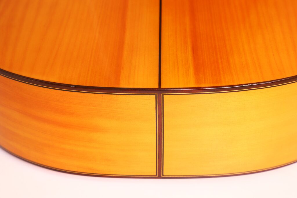 Nahaufnahme der Rückseite und der Seite einer Flamenco-Gitarre von Antonio Bernal aus dem Jahr 2009, die das glatte, polierte helle Holz mit dunklen Zierleisten, sichtbaren Maserungen und Details entlang der Rundung und der Naht des Instruments hervorhebt. Nahaufnahme der Rückseite und der Seite einer Flamenco-Gitarre von Antonio Bernal aus dem Jahr 2009, die das glatte, polierte helle Holz mit dunklen Zierleisten, sichtbaren Maserungen und Details entlang der Rundung und der Naht des Instruments hervorhebt.