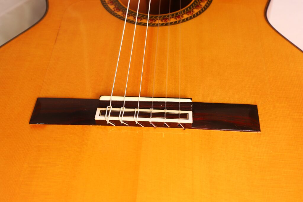 Nahaufnahme des Stegs und der Saiten einer Flamenco-Gitarre von Antonio Bernal aus dem Jahr 2009, die die Maserung des Holzes und die feine Handwerkskunst hervorhebt. Nahaufnahme des Stegs und der Saiten einer Flamenco-Gitarre von Antonio Bernal aus dem Jahr 2009, die die Maserung des Holzes und die feine Handwerkskunst hervorhebt.