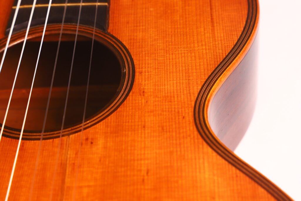 Nahaufnahme des Korpus der Romantikgitarre Markneukirchen 1880, die die detaillierte Holzstruktur, die Saiten und die geschwungene Kante am Schallloch zeigt, die die Handwerkskunst und die warmen Holztöne hervorhebt. Nahaufnahme des Korpus der Romantikgitarre Markneukirchen 1880, die die detaillierte Holzstruktur, die Saiten und die geschwungene Kante am Schallloch zeigt, die die Handwerkskunst und die warmen Holztöne hervorhebt.