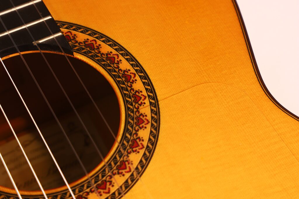 Nahaufnahme einer Flamenco-Gitarre von Antonio Bernal aus dem Jahr 2009, die das Schallloch mit seiner detailreichen Rosette sowie einen Teil der Saiten und den Holzkorpus der Gitarre zeigt. Nahaufnahme einer Flamenco-Gitarre von Antonio Bernal aus dem Jahr 2009, die das Schallloch mit seiner detailreichen Rosette sowie einen Teil der Saiten und den Holzkorpus der Gitarre zeigt.