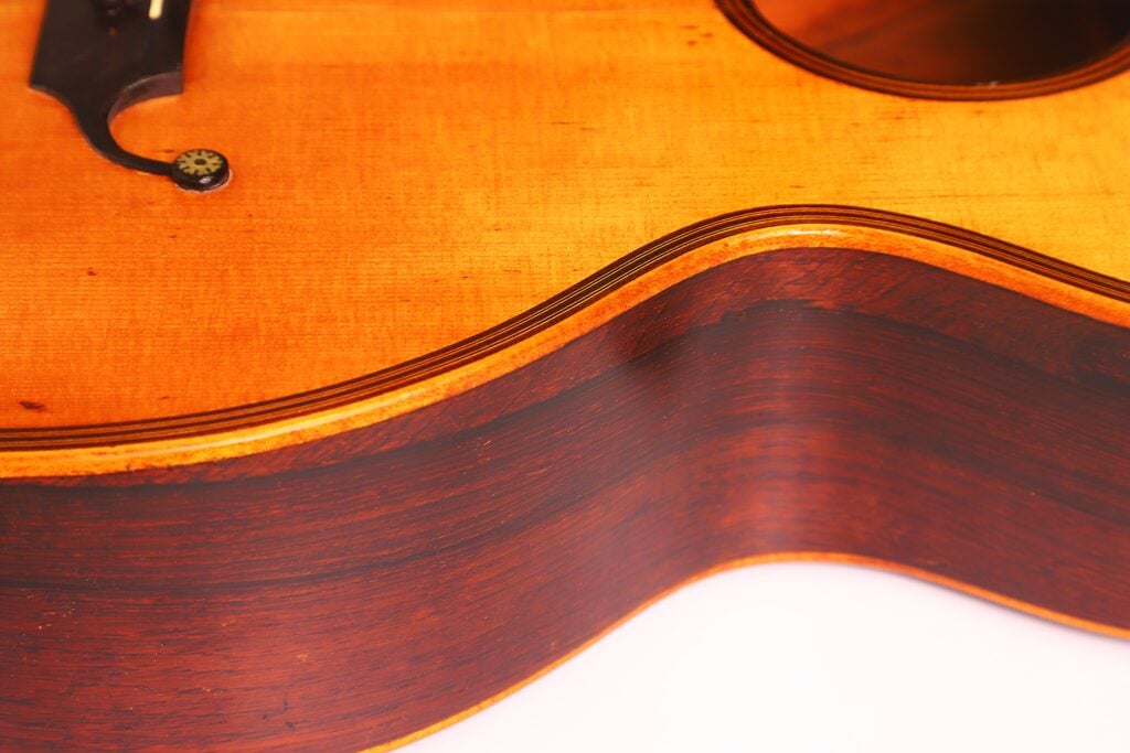 Nahaufnahme des Holzkorpus der Markneukirchen 1880 Romantikgitarre, die die sanfte Wölbung, die sichtbaren Maserungen und das teilweise Schallloch zeigt. Die Gitarre hat eine natürliche Lackierung mit kontrastierenden dunkelbraunen Zargen und einer helleren Decke. Nahaufnahme des Holzkorpus der Markneukirchen 1880 Romantikgitarre, die die sanfte Wölbung, die sichtbaren Maserungen und das teilweise Schallloch zeigt. Die Gitarre hat eine natürliche Lackierung mit kontrastierenden dunkelbraunen Zargen und einer helleren Decke.