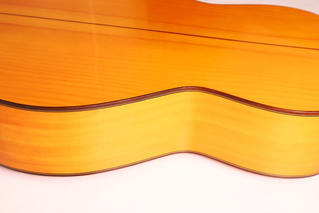 Nahaufnahme der Seite und Rückseite einer Flamenco-Gitarre von Antonio Bernal aus dem Jahr 2009, die die glatte, glänzende Holzoberfläche mit hellbrauner Lackierung und die elegant geschwungene Form hervorhebt. Nahaufnahme der Seite und Rückseite einer Flamenco-Gitarre von Antonio Bernal aus dem Jahr 2009, die die glatte, glänzende Holzoberfläche mit hellbrauner Lackierung und die elegant geschwungene Form hervorhebt.
