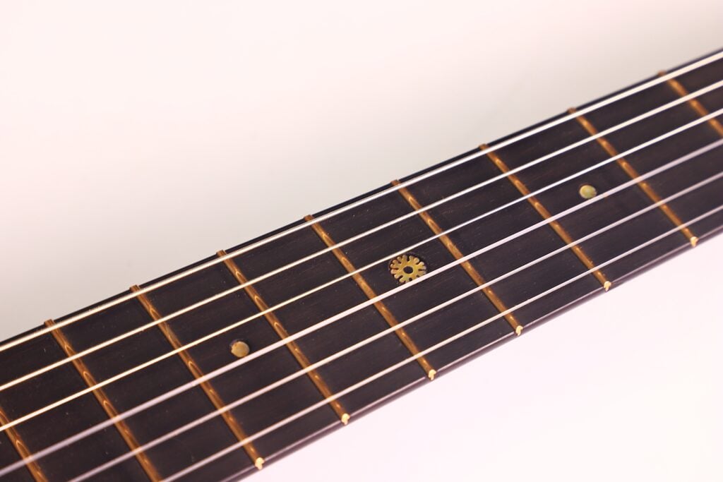 Eine Nahaufnahme des Griffbretts der Markneukirchen 1880 Romantikgitarre mit Metallbünden, Punkteinlagen und einer dekorativen zahnradförmigen Einlage, dargestellt vor einem weißen Hintergrund. Eine Nahaufnahme des Griffbretts der Markneukirchen 1880 Romantikgitarre mit Metallbünden, Punkteinlagen und einer dekorativen zahnradförmigen Einlage, dargestellt vor einem weißen Hintergrund.