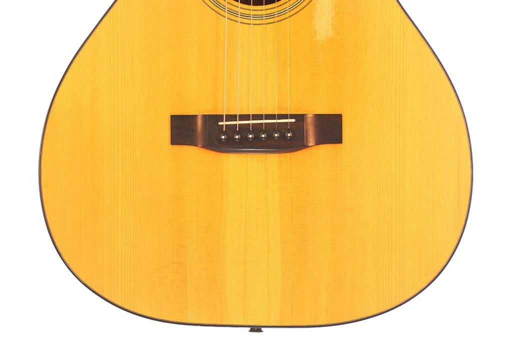 Nahaufnahme der Rozawood 000 12-18-HM Akustikgitarre, die den Holzsteg, die Saiten und die helle Holzmaserung in der unteren Hälfte des Korpus hervorhebt. Nahaufnahme der Rozawood 000 12-18-HM Akustikgitarre, die den Holzsteg, die Saiten und die helle Holzmaserung in der unteren Hälfte des Korpus hervorhebt.