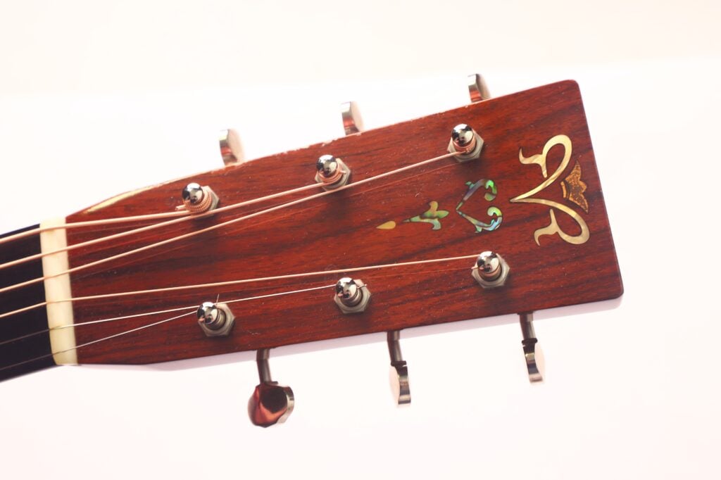 Nahaufnahme der hölzernen Kopfplatte der Rozawood 000 12-18-HM Akustikgitarre mit silbernen Stimmwirbeln, Ziereinlage und sechs Saiten vor einem schlichten weißen Hintergrund. Nahaufnahme der hölzernen Kopfplatte der Rozawood 000 12-18-HM Akustikgitarre mit silbernen Stimmwirbeln, Ziereinlage und sechs Saiten vor einem schlichten weißen Hintergrund.