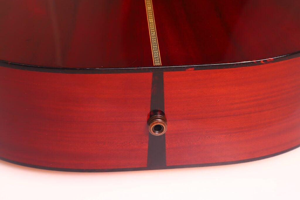 Nahaufnahme der Unterseite der Rozawood 000 12-18-HM Akustikgitarre mit glänzendem rotbraunem Finish, dekorativer Einlage und Metalleingangsbuchse für den elektrischen Tonabnehmer. Nahaufnahme der Unterseite der Rozawood 000 12-18-HM Akustikgitarre mit glänzendem rotbraunem Finish, dekorativer Einlage und Metalleingangsbuchse für den elektrischen Tonabnehmer.