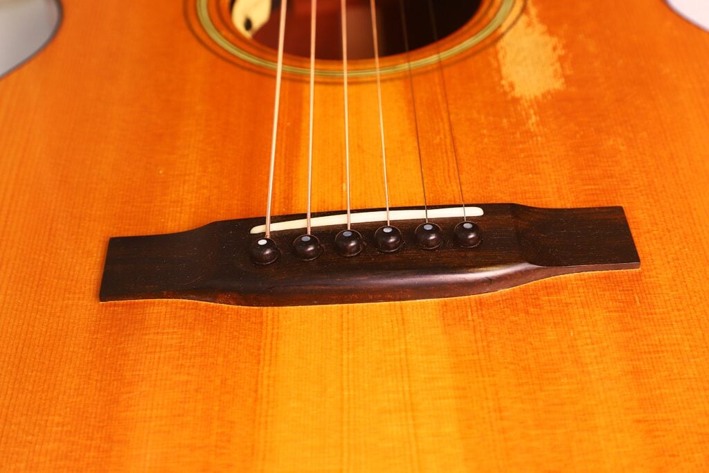 Nahaufnahme des Stegs und der Saiten der Rozawood 000 12-18-HM Akustik Gitarre, die die Holzmaserung und die detaillierte Verarbeitung des Korpus und der Stegstifte hervorhebt. Nahaufnahme des Stegs und der Saiten der Rozawood 000 12-18-HM Akustik Gitarre, die die Holzmaserung und die detaillierte Verarbeitung des Korpus und der Stegstifte hervorhebt.