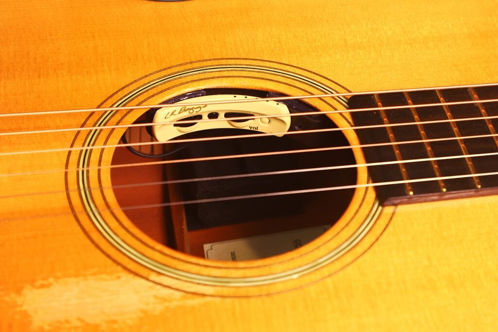 Nahaufnahme des Schalllochs einer Rozawood 000 12-18-HM Akustik Gitarre, die einen Tonabnehmer unter den Saiten zeigt. Sichtbar sind die Holzmaserung, ein Teil des Griffbretts und das Markenetikett im Inneren des Gitarrenkorpus. Nahaufnahme des Schalllochs einer Rozawood 000 12-18-HM Akustik Gitarre, die einen Tonabnehmer unter den Saiten zeigt. Sichtbar sind die Holzmaserung, ein Teil des Griffbretts und das Markenetikett im Inneren des Gitarrenkorpus.