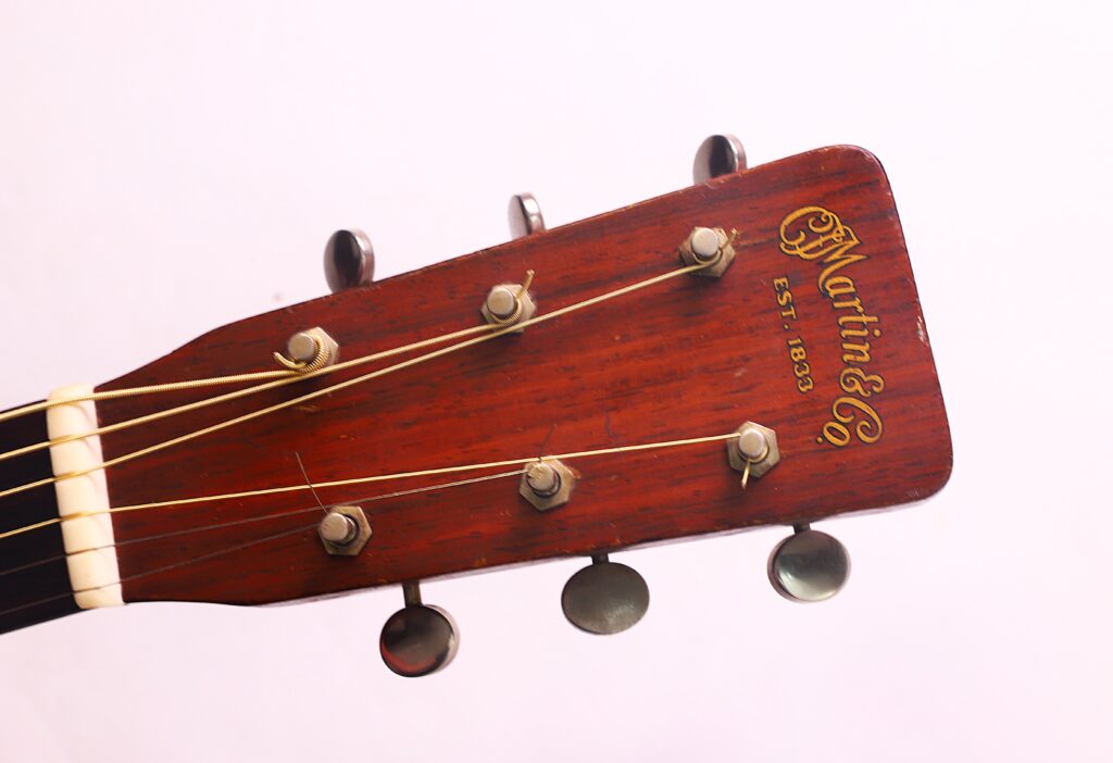 Nahaufnahme des Kopfes der Martin 00-18 Akustikgitarre aus dem Jahr 1954 mit sechs Stimmwirbeln und dem goldenen „Martin & Co. Est. 1833“-Logo vor einem schlichten, hellen Hintergrund. Nahaufnahme des Kopfes der Martin 00-18 Akustikgitarre aus dem Jahr 1954 mit sechs Stimmwirbeln und dem goldenen „Martin & Co. Est. 1833“-Logo vor einem schlichten, hellen Hintergrund.