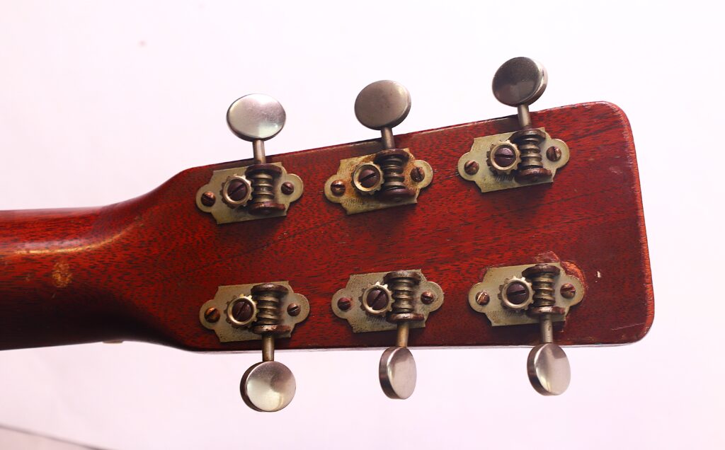 Nahaufnahme der Rückseite des Kopfes einer Martin 00-18 Akustikgitarre aus dem Jahr 1954, mit sechs Metallstimmwirbeln auf einer rotbraunen Holzoberfläche vor einem schlichten Hintergrund. Nahaufnahme der Rückseite des Kopfes einer Martin 00-18 Akustikgitarre aus dem Jahr 1954, mit sechs Metallstimmwirbeln auf einer rotbraunen Holzoberfläche vor einem schlichten Hintergrund.