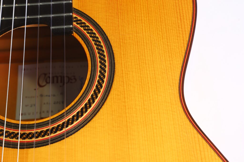 Nahaufnahme des Korpus der Flamenco-Gitarre „Camps Primera A 2018“ mit Blick auf das Schallloch, die Saiten, die Zierrosette, die Holzmaserung und einen Teil des Innenetiketts. Nahaufnahme des Korpus der Flamenco-Gitarre „Camps Primera A 2018“ mit Blick auf das Schallloch, die Saiten, die Zierrosette, die Holzmaserung und einen Teil des Innenetiketts.