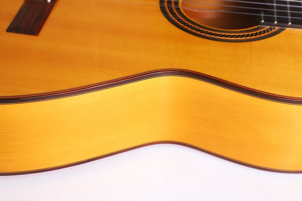 Nahaufnahme des Korpus der Flamenco-Gitarre „Camps Primera A 2018“, die ihre glatte, geschwungene Seite sowie einen Teil der Saiten und das Schallloch vor einem weißen Hintergrund hervorhebt. Nahaufnahme des Korpus der Flamenco-Gitarre „Camps Primera A 2018“, die ihre glatte, geschwungene Seite sowie einen Teil der Saiten und das Schallloch vor einem weißen Hintergrund hervorhebt.