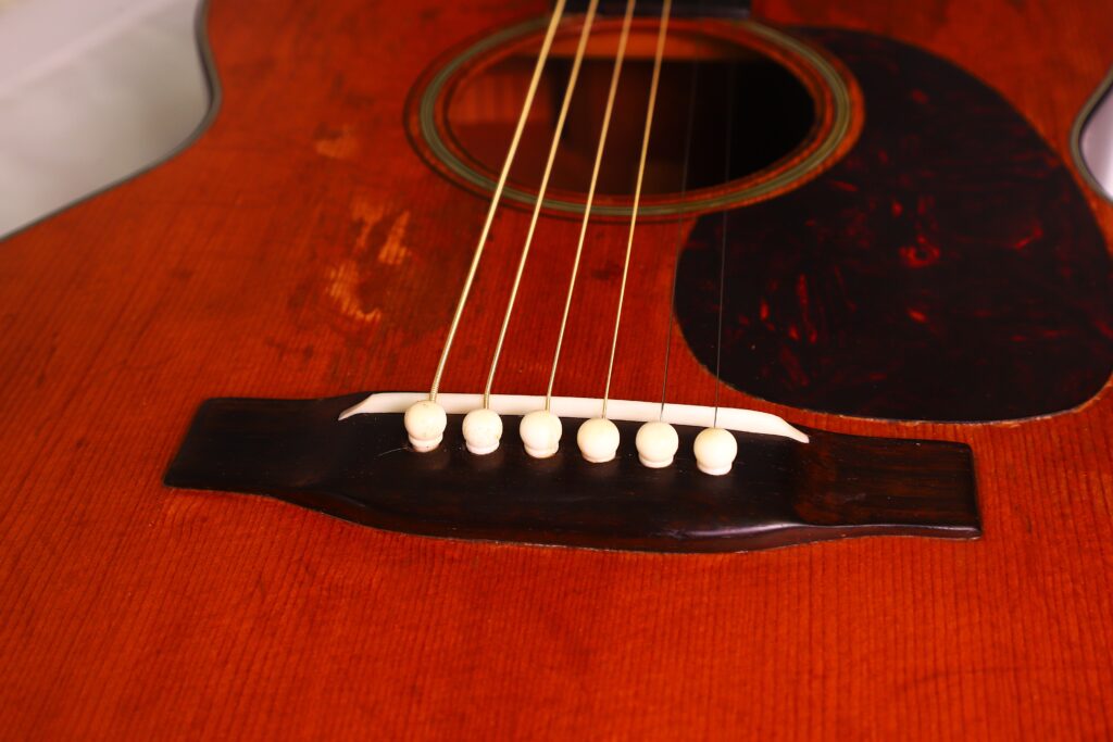 Nahaufnahme des Holzkörpers einer Martin 00-18 Akustikgitarre aus dem Jahr 1954, auf der die sechs Saiten, der Steg mit weißen Stiften, die rotbraune Lackierung und das dunkle Schlagbrett zu sehen sind. Nahaufnahme des Holzkörpers einer Martin 00-18 Akustikgitarre aus dem Jahr 1954, auf der die sechs Saiten, der Steg mit weißen Stiften, die rotbraune Lackierung und das dunkle Schlagbrett zu sehen sind.