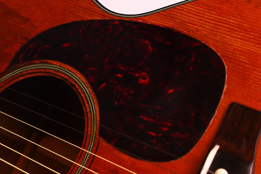 Nahaufnahme einer Martin 00-18 Akustikgitarre aus dem Jahr 1954, bei der das Schallloch, die Saiten und das dunkle, im Schildpatt-Muster gehaltene Schlagbrett auf dem Holzkorpus besonders hervorgehoben sind. Nahaufnahme einer Martin 00-18 Akustikgitarre aus dem Jahr 1954, bei der das Schallloch, die Saiten und das dunkle, im Schildpatt-Muster gehaltene Schlagbrett auf dem Holzkorpus besonders hervorgehoben sind.