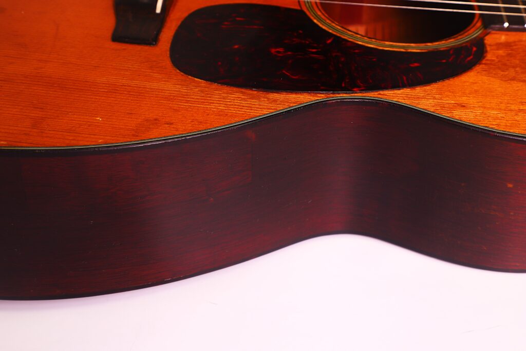 Nahaufnahme des unteren Teils der Martin 00-18 Akustikgitarre aus dem Jahr 1954, bei der die Holzmaserung, die Saiten, das Schlagbrett und die geschwungene Seite vor einem weißen Hintergrund hervorgehoben werden. Nahaufnahme des unteren Teils der Martin 00-18 Akustikgitarre aus dem Jahr 1954, bei der die Holzmaserung, die Saiten, das Schlagbrett und die geschwungene Seite vor einem weißen Hintergrund hervorgehoben werden.
