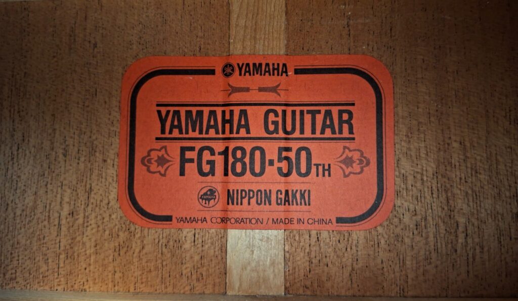 Ein orangefarbenes Etikett auf Holz lautet: Yamaha FG-180 50th Anniversary Akustik Gitarre, zeigt das Yamaha-Logo und die Angabe Yamaha Corporation / Made in China. Ein orangefarbenes Etikett auf Holz lautet: Yamaha FG-180 50th Anniversary Akustik Gitarre, zeigt das Yamaha-Logo und die Angabe Yamaha Corporation / Made in China.