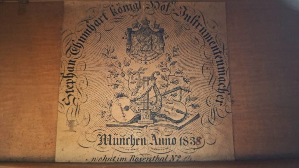 Ein Vintage-Etikett für die Stephan-Thumhart-Romantikgitarre von 1838 zieren kunstvolle deutsche Schriftzüge, ein Wappen und Abbildungen von Musikinstrumenten mit der Aufschrift: „Stephan Thumhart, königlicher Hof-Instrumentenmacher, München, Anno 1838“. Ein Vintage-Etikett für die Stephan-Thumhart-Romantikgitarre von 1838 zieren kunstvolle deutsche Schriftzüge, ein Wappen und Abbildungen von Musikinstrumenten mit der Aufschrift: „Stephan Thumhart, königlicher Hof-Instrumentenmacher, München, Anno 1838“.
