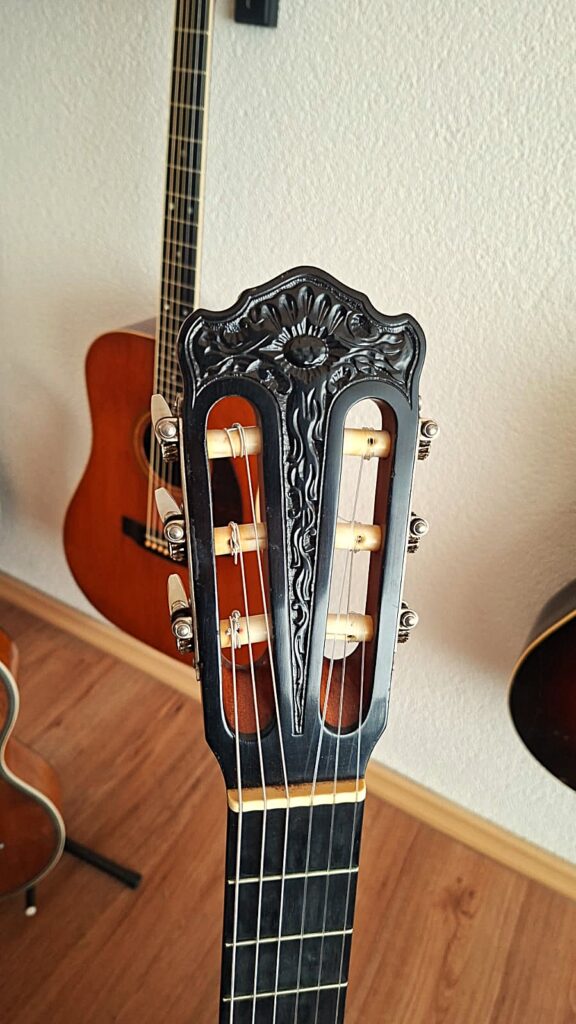 Nahaufnahme des kunstvoll verzierten Kopfes einer „Klassischen Gitarre“ von Joachim Schneider aus dem Jahr 1977 mit aufwendigen Blumenverzierungen; im Hintergrund sind zwei weitere Akustikgitarren zu sehen, die alle auf Ständern auf einem Holzboden vor einer weißen, strukturierten Wand stehen. Nahaufnahme des kunstvoll verzierten Kopfes einer „Klassischen Gitarre“ von Joachim Schneider aus dem Jahr 1977 mit aufwendigen Blumenverzierungen; im Hintergrund sind zwei weitere Akustikgitarren zu sehen, die alle auf Ständern auf einem Holzboden vor einer weißen, strukturierten Wand stehen.