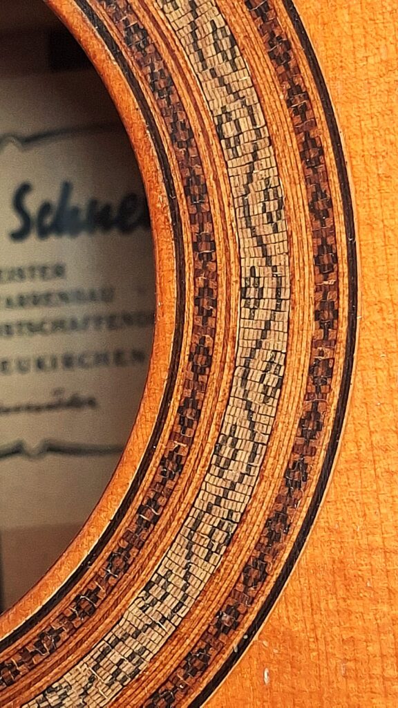 Nahaufnahme des Schalllochs der „Joachim Schneider 1977 Klassische Gitarre“ mit detaillierten Mosaik-Einlagen am Rand. Durch das Schallloch ist im Inneren des Korpus ein Teil des Etiketts der Gitarre mit unscharfem Text zu sehen. Nahaufnahme des Schalllochs der „Joachim Schneider 1977 Klassische Gitarre“ mit detaillierten Mosaik-Einlagen am Rand. Durch das Schallloch ist im Inneren des Korpus ein Teil des Etiketts der Gitarre mit unscharfem Text zu sehen.