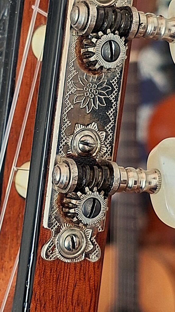 Nahaufnahme der Stimmwirbel und der verzierten Metallplatte am Kopf einer „Klassischen Gitarre“ von Joachim Schneider aus dem Jahr 1977, die die filigranen Blumengravuren und die Details der Holzmaserung hervorhebt. Nahaufnahme der Stimmwirbel und der verzierten Metallplatte am Kopf einer „Klassischen Gitarre“ von Joachim Schneider aus dem Jahr 1977, die die filigranen Blumengravuren und die Details der Holzmaserung hervorhebt.