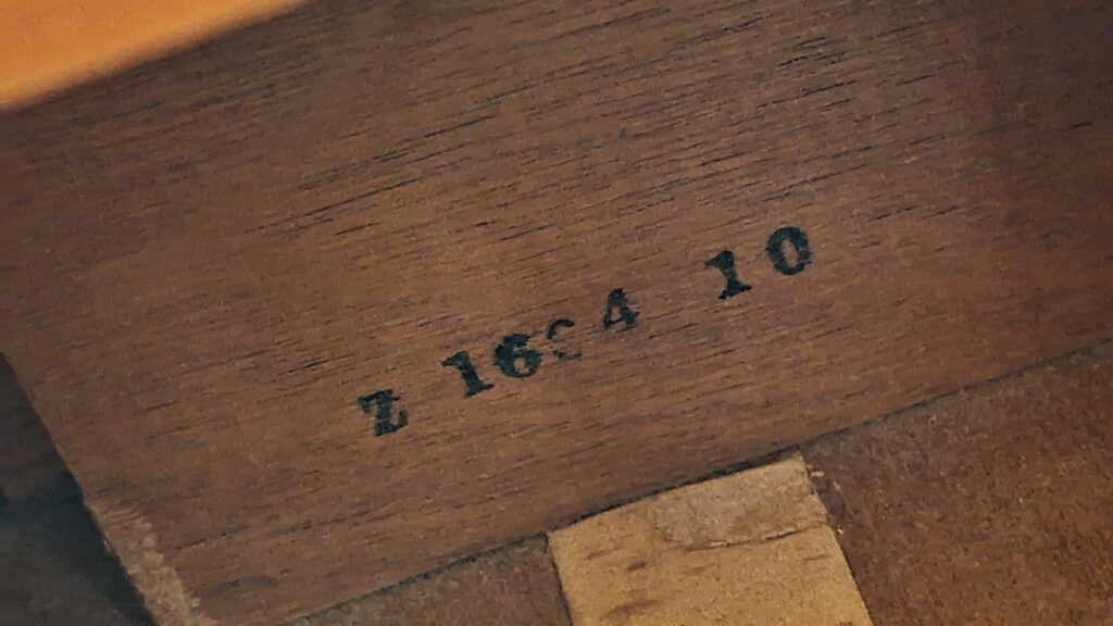 Eine Nahaufnahme der Holzoberfläche der Gibson J-50 Akustikgitarre aus dem Jahr 1952 zeigt den schwarz eingeprägten Text „E 16 4 10“ sowie einen Streifen aus hellerem Material – möglicherweise Stoff oder Papier – entlang der Unterkante. Eine Nahaufnahme der Holzoberfläche der Gibson J-50 Akustikgitarre aus dem Jahr 1952 zeigt den schwarz eingeprägten Text „E 16 4 10“ sowie einen Streifen aus hellerem Material – möglicherweise Stoff oder Papier – entlang der Unterkante.