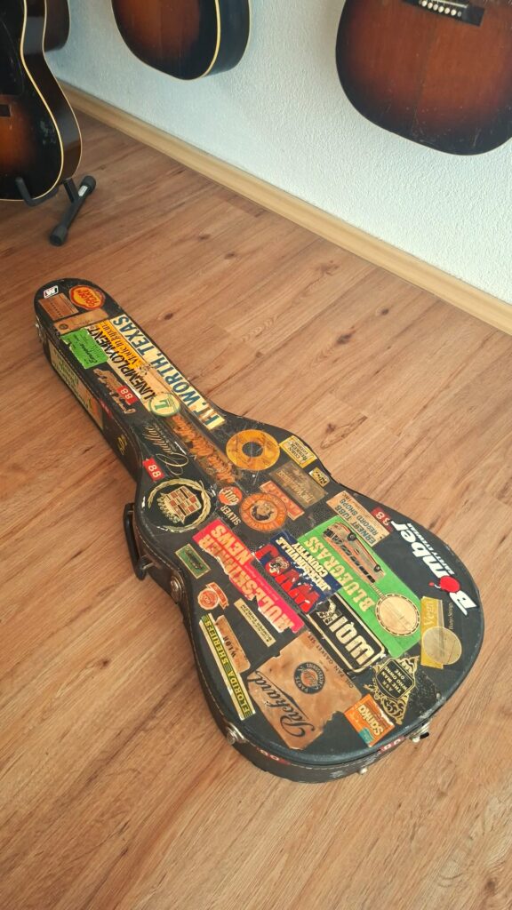 Ein schwarzer Gitarrenkoffer, der mit bunten Aufklebern beklebt ist, steht auf einem Holzboden, während im Hintergrund mehrere Martin 00-18 Akustikgitarren aus dem Jahr 1954 an der weißen Wand hängen. Ein schwarzer Gitarrenkoffer, der mit bunten Aufklebern beklebt ist, steht auf einem Holzboden, während im Hintergrund mehrere Martin 00-18 Akustikgitarren aus dem Jahr 1954 an der weißen Wand hängen.