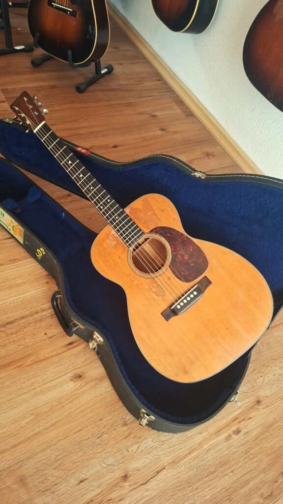 Eine Martin 00-18 Akustikgitarre aus dem Jahr 1954 mit naturbelassener Holzoberfläche liegt in einem offenen, mit blauem Samt ausgekleideten Hartschalenkoffer auf einem Holzboden, während hinter ihr mehrere andere Gitarren an der Wand hängen. Eine Martin 00-18 Akustikgitarre aus dem Jahr 1954 mit naturbelassener Holzoberfläche liegt in einem offenen, mit blauem Samt ausgekleideten Hartschalenkoffer auf einem Holzboden, während hinter ihr mehrere andere Gitarren an der Wand hängen.