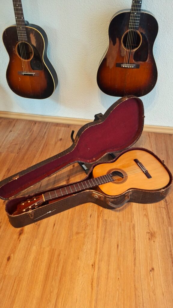 Eine klassische Gitarre von Jose Ramirez I aus dem Jahr 1921 steht in einem offenen Hartschalenkoffer mit rotem Innenfutter auf einem Holzboden, während im Hintergrund zwei Vintage-Akustikgitarren an einer weißen Wand hängen. Eine klassische Gitarre von Jose Ramirez I aus dem Jahr 1921 steht in einem offenen Hartschalenkoffer mit rotem Innenfutter auf einem Holzboden, während im Hintergrund zwei Vintage-Akustikgitarren an einer weißen Wand hängen.