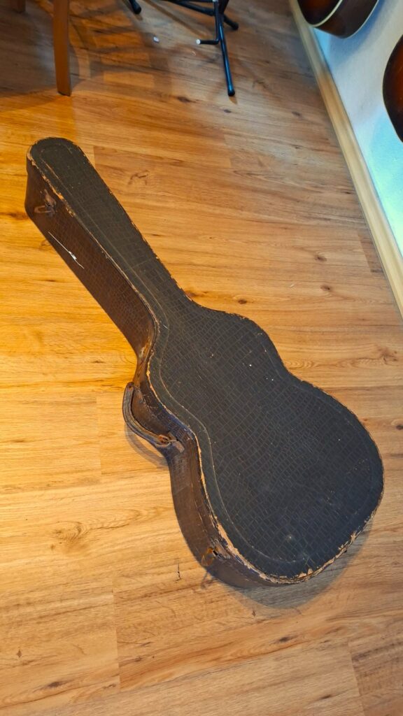 In einem abgenutzten, schwarzen Vintage-Koffer mit Kratzern und ausgefransten Kanten befindet sich eine „Jose Ramirez I 1921 Klassische Gitarre“, die geschlossen auf einem Holzboden in der Nähe einer Wand liegt, an der weitere Gitarren ausgestellt sind. In einem abgenutzten, schwarzen Vintage-Koffer mit Kratzern und ausgefransten Kanten befindet sich eine „Jose Ramirez I 1921 Klassische Gitarre“, die geschlossen auf einem Holzboden in der Nähe einer Wand liegt, an der weitere Gitarren ausgestellt sind.