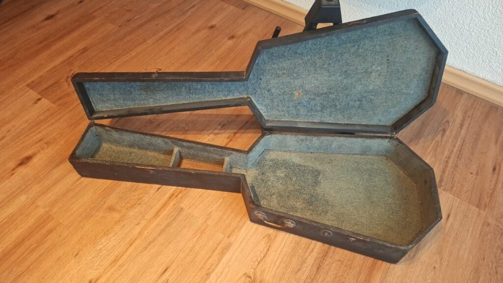 Ein abgenutzter Holzkoffer für eine Romantikgitarre aus der Zeit um 1820 mit blauem Innenfutter und Zubehörfächern steht offen und leer auf einem Holzboden neben einer weißen Wand. Ein abgenutzter Holzkoffer für eine Romantikgitarre aus der Zeit um 1820 mit blauem Innenfutter und Zubehörfächern steht offen und leer auf einem Holzboden neben einer weißen Wand.