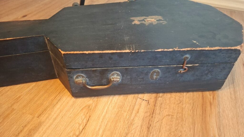 Der „Holzkoffer ~1820 Romantikgitarre“ ist ein schwarzer, sargförmiger Holzkasten mit einem Metallgriff und einem Verschluss, dessen abgeplatzte Kanten und Kratzer von seinem Alter und seiner häufigen Nutzung zeugen. Der „Holzkoffer ~1820 Romantikgitarre“ ist ein schwarzer, sargförmiger Holzkasten mit einem Metallgriff und einem Verschluss, dessen abgeplatzte Kanten und Kratzer von seinem Alter und seiner häufigen Nutzung zeugen.