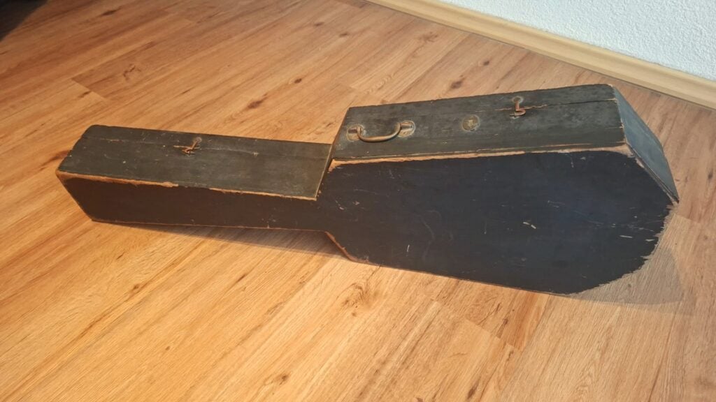 Ein abgenutzter, schwarzer Vintage-Holzkoffer ~1820 für eine Romantikgitarre mit rechteckigem Griff steht auf einem hellen Holzboden neben einer weißen Wand; er ist für eine Gitarre oder ein ähnliches Instrument gedacht. Ein abgenutzter, schwarzer Vintage-Holzkoffer ~1820 für eine Romantikgitarre mit rechteckigem Griff steht auf einem hellen Holzboden neben einer weißen Wand; er ist für eine Gitarre oder ein ähnliches Instrument gedacht.