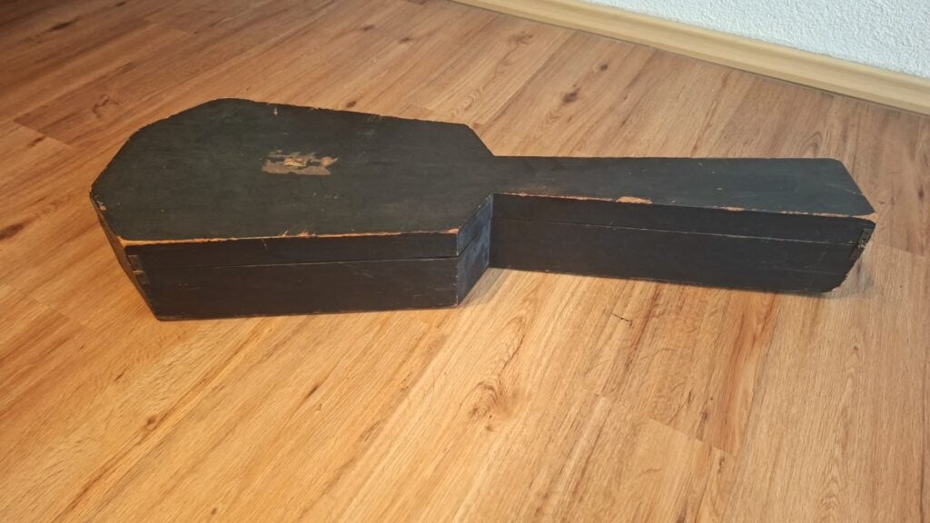 Ein Holzkoffer mit einer Romantikgitarre aus der Zeit um 1820, schwarz mit sichtbaren Kratzern und abgenutzten Kanten, steht auf einem Holzboden in der Nähe einer weißen Wand und einer Holzfußleiste. Ein Holzkoffer mit einer Romantikgitarre aus der Zeit um 1820, schwarz mit sichtbaren Kratzern und abgenutzten Kanten, steht auf einem Holzboden in der Nähe einer weißen Wand und einer Holzfußleiste.