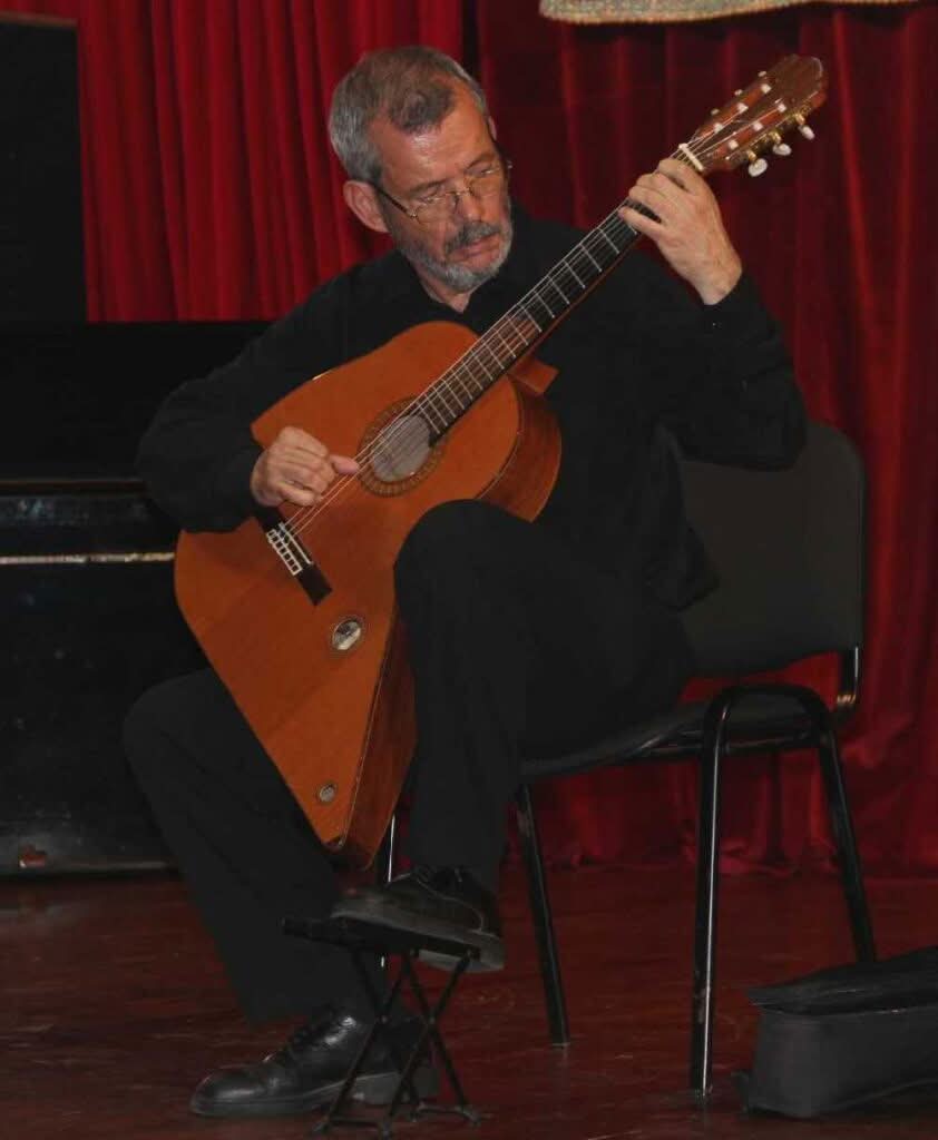 Ein Mann mit grauen Haaren und Brille sitzt auf einem Stuhl und spielt auf der Bühne die „Gran Klassische Gitarre“ von Raimundo y Aparicio Corujo, hinter ihm sind rote Vorhänge zu sehen. Ganz in Schwarz gekleidet, scheint er sich ganz auf das Instrument zu konzentrieren. Ein Mann mit grauen Haaren und Brille sitzt auf einem Stuhl und spielt auf der Bühne die „Gran Klassische Gitarre“ von Raimundo y Aparicio Corujo, hinter ihm sind rote Vorhänge zu sehen. Ganz in Schwarz gekleidet, scheint er sich ganz auf das Instrument zu konzentrieren.